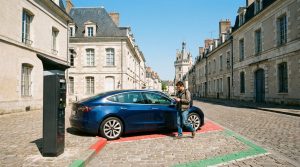 Un homme près d'une voiture Tesla bleue et un horodateur dans une rue pavée de Blois. Bâtiments historiques et une tour sous un ciel bleu.