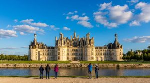 Château de Chambord, architecture Renaissance imposante, sous ciel bleu. Cinq visiteurs l'admirent depuis le bord de l'eau.